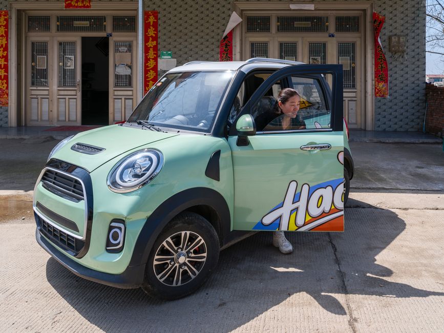 Young woman exits vibrant electric car parked in an urban area, surrounded by cultural decorations.